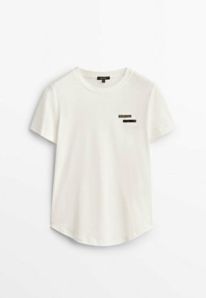 T-shirt blanc à manches courtes avec col rond et deux petites bandes horizontales noires et dorées pailletées sur le côté gauche de la poitrine.