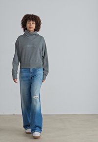 Grauer kurzer Sweatshirt mit hohem Kragen und Logodetail, kombiniert mit weiten blauen Jeans mit distressed Akzenten. Weiße Sneaker runden den Look ab.