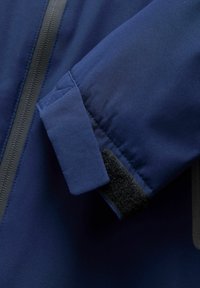 Marineblauwe jas met een zachte, gladde textuur, voorzien van een klittenbandsluiting aan de manchetten en een contrasterende zwarte ritssluiting.