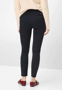 Zwarte nauwsluitende broek met een gladde textuur, twee achterzakken en een taille detail. Gecombineerd met bordeauxrode platte schoenen.