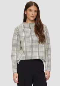 Lange mouwen, crèmekleurig top met een zwart rasterpatroon. Heeft een hoge kraag en een losse pasvorm. Draagt met een zwarte broek.