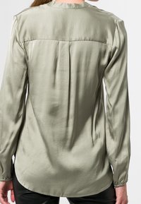 Blouse vert olive à manches longues avec une texture lisse, un col rond, des poignets froncés et un pli décoratif au centre du dos.