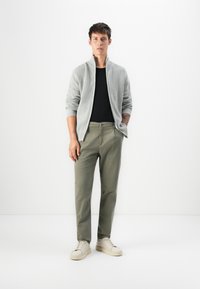 Unge mann står mot en hvit bakgrunn iført en lys grå zip-up genser, svart skjorte, olivengrønne bukser og hvite sneakers.