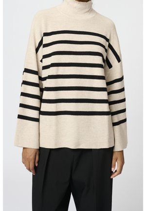 Personne portant un pull à col roulé beige avec des rayures noires horizontales et des manches larges, associé à un pantalon noir ample.