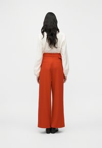 Παντελόνια με φαρδιά μπατζάκια και ψηλή μέση από κόκκινο ύφασμα με λεία υφή, διακοσμημένα με ζώνη και δύο πίσω τσέπες. Ορατά μαύρα παπούτσια.