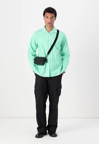 Camisa verde menta com riscas verticais e botões, combinada com calças cargo pretas e uma pequena mala de ombro preta, usada por um modelo masculino.
