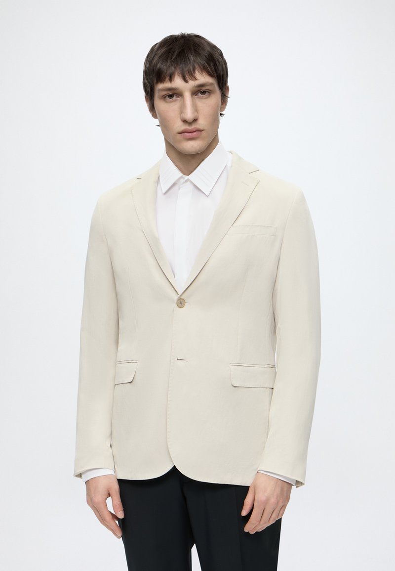 Jeune homme portant un blazer beige à un seul bouton et une chemise blanche, debout devant un fond clair uni.