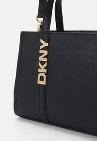 Juoda struktūruota rankinė, pagaminta iš tekstūrinės sintetinės medžiagos, su auksinėmis detalėmis ir "DKNY" logotipu blokinėmis raidėmis bei dviem rankenomis.