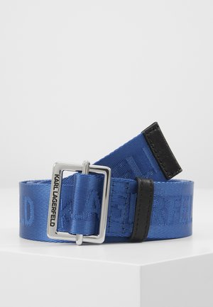 Ceinture en tissu bleu avec accents en cuir noir et boucle rectangulaire argentée gravée de "Karl Lagerfeld" sur une surface blanche.