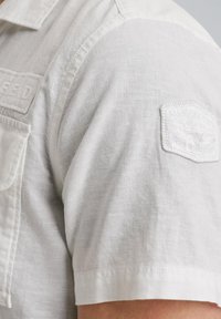 Camisa de manga corta en lino beis claro. Presenta una superficie texturizada, un parche en la manga izquierda con un logo en relieve y un cuello abotonado.