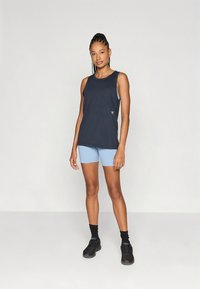 Marineblauw mouwloos tanktop met een subtiel logo, gecombineerd met lichtblauwe shorts. Model draagt zwarte enkelsokken en sportschoenen.