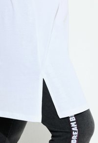 Witte katoenen T-shirt met zijslitsen, gecombineerd met donkergrijze leggings met een verticale witte logo-print langs de zijkant.