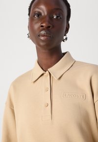 Polo beige con una texture morbida, caratterizzato da un colletto classico, una doppia fila di bottoni e il logo "LACOSTE" impresso sul petto.