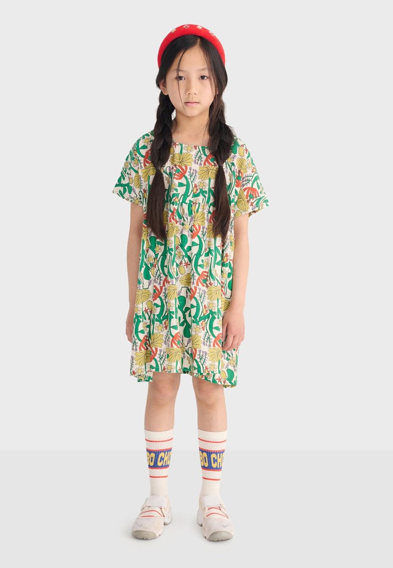 Fille aux longs cheveux tressés, vêtue d'une robe fleurie, d'un serre-tête rouge, de chaussettes blanches avec du texte bleu et jaune, et de baskets blanches, debout devant un fond blanc.