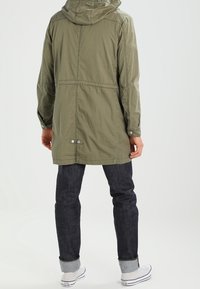 Parka à capuche vert olive, longue avec une coupe décontractée, présentant un détail de couture, des boutons-pression et des poignets élastiques, portée sur un jean foncé.