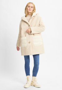 Cappotto beige texturizzato con cappuccio, dotato di due grandi tasche frontali imbottite e chiusure a bottoni. Abbinato a jeans blu e stivali in pelle beige con lacci.