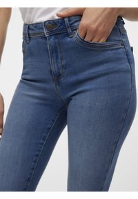 Gros plan d'une personne portant un jean en denim bleu, avec une main dans la poche avant, sur un fond clair uni.