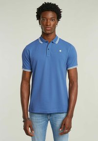 Blå pikétröja med vit krage och ärmsprund, korta ärmar och en tvåknappsknäppning. Har en liten logotyp på bröstet.