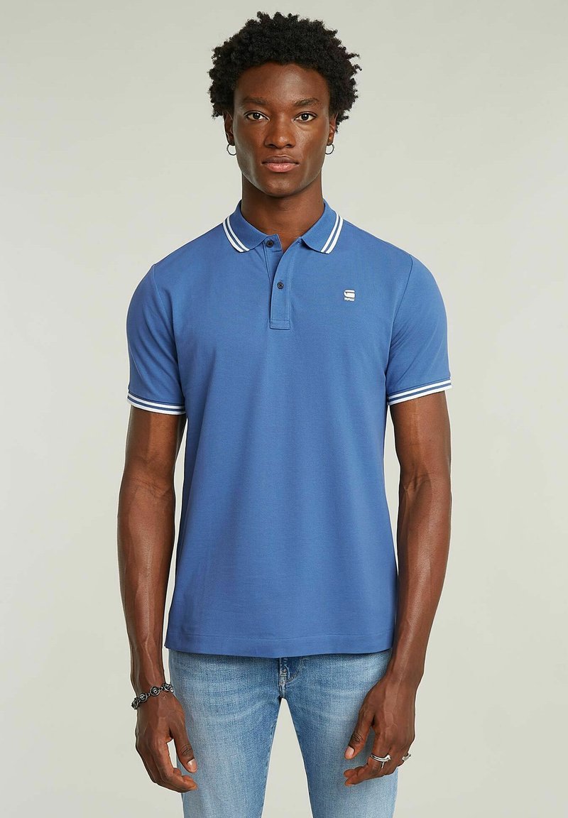 Blå pikétröja med vit krage och ärmsprund, korta ärmar och en tvåknappsknäppning. Har en liten logotyp på bröstet.