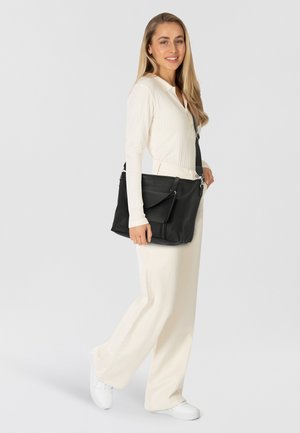 Borsa in pelle nera con finitura testurizzata, dotata di chiusura con zip, tracolla regolabile e tasca frontale. Modello che indossa una maglia bianca a maniche lunghe.
