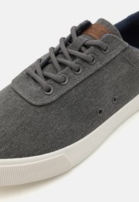 Gray canvas sneaker with flat laces, rubber sole, and a brown leather accent on the tongue. Features metal eyelets and a textured surface.