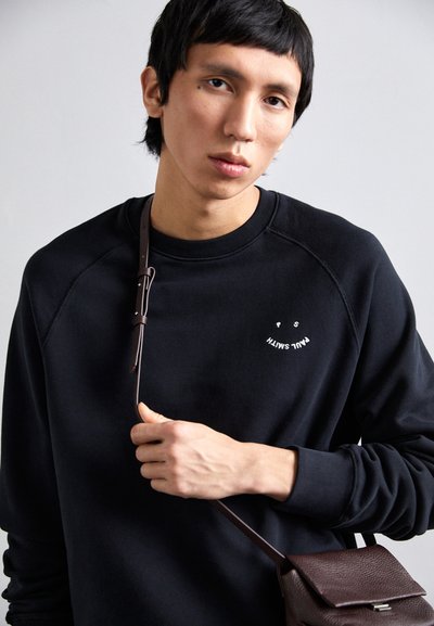 Svart crewneck genser med raglanermer, med et lite hvitt grafisk trykk på brystet. Personen holder et brunt lærreim fra en bag.
