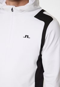 Vit hoodie med en kvartdragkedja, med svarta sidodetaljer och en liten logotyp. Tyget verkar slätt med en subtil lyster.