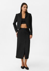 Zwart op maat gemaakte cropped blazer met bijpassende hoge taille rok; gladde textuur, enkele knoop en splitdetail aan de voorkant; gecombineerd met zwarte flats.