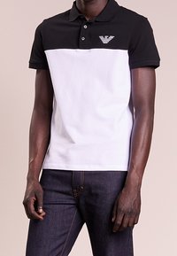 Svart och vit polo med skjortkrage, med en liten logotyp på bröstet. Skjortan har korta ärmar och en kontrasterande fåll.