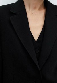 Gros plan sur une personne portant un blazer noir par-dessus un haut noir à encolure en V profonde avec des détails de boutons.