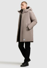 Cappotto imbottito beige con colletto alto, chiusura con zip e due tasche laterali. Presenta un'etichetta con logo all'orlo e un design con cappuccio.