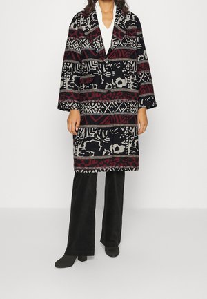 Femme portant un manteau à motifs noir, rouge et gris longueur genou sur un haut blanc et un pantalon large noir avec des chaussures à talons noires.