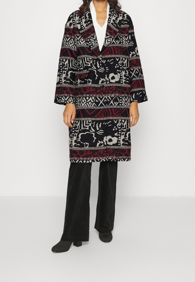 Femme portant un manteau à motifs noir, rouge et gris longueur genou sur un haut blanc et un pantalon large noir avec des chaussures à talons noires.