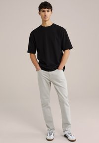 Zwart t-shirt met korte mouwen en ronde hals, gecombineerd met lichtgrijze jeans en zwart-witte sneakers op een neutrale achtergrond.
