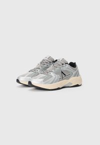 Zapatillas deportivas plateadas con paneles de malla, superposiciones texturizadas, acentos negros y suelas beige. Presentan cierre con cordones y un interior acolchado.