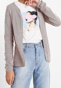 Ljusrosa cardigan med ett texturerat geometriskt mönster, kombinerad med en vit t-shirt med grafik och högmidjade ljusblå jeans.