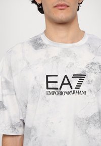 Balta trumpų rankovių marškinėliai su marmuro tekstūra, su juodu EA7 Emporio Armani logotipu ant krūtinės. Apvali iškirptė, laisvas siluetas.