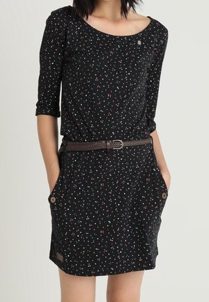 Frau in einem schwarzen Kleid mit kleinen, bunten Punkten, dreiviertel Ärmeln, seitlichen Taschen mit Knöpfen und einem braunen geflochtenen Gürtel.