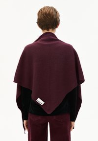 Poncho en mélange de laine bordeaux avec une forme triangulaire, une texture lisse et une étiquette de marque visible sur le bord inférieur.