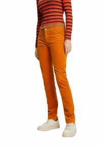 Pantalon en velours côtelé orange à coupe slim, avec fermeture à boutons et poches avant, associé à des baskets blanches et un haut à manches longues rayé.