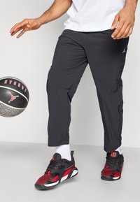 Μαύρο αθλητικό παντελόνι με στενή εφαρμογή, συνδυασμένο με μαύρα και κόκκινα αθλητικά παπούτσια, με διακριτικά στοιχεία δικτύου, και μια μπάλα μπάσκετ σε κίνηση.