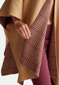 Béžový vlněný pončo s podšívkou ve vzoru houndstooth v odstínech burgundské a smetanové. Má volný střih a jemnou texturu.