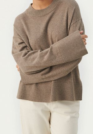 Personne portant un pull en grosse maille marron oversize avec poignets et ourlet côtelés, associé à un pantalon beige clair, bras croisés devant.