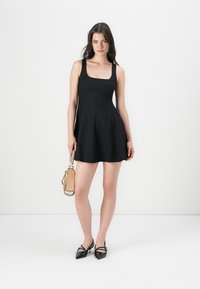 Zwart mouwloos jurk met een aansluitend lijfje en een uitlopende rok. Model houdt een kleine beige handtas vast en draagt zwarte puntige ballerina's. Gladde textuur.