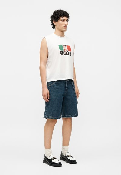 Άνδρας με λευκό αμάνικο γραφικό μπλουζάκι, σκούρα τζιν σόρτς, λευκές κάλτσες και ασπρόμαυρα μοκασίνια, στέκεται μπροστά σε απλό φόντο.