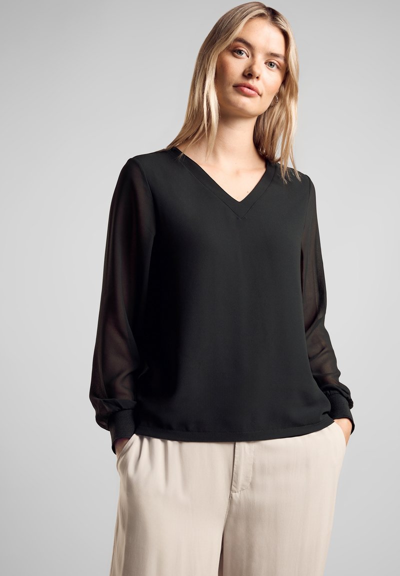 Blusa nera con scollatura a V, maniche lunghe e trasparenti, e una consistenza liscia. Abbinata a pantaloni beige a vita alta, con dettagli plissettati.