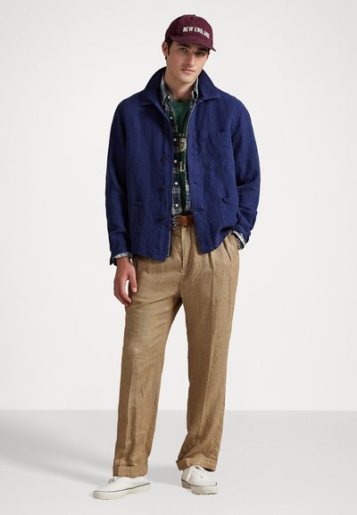 Navy blue linen jacket over a green shirt and plaid flannel, paired with brown checked trousers and white sneakers, topped with a maroon cap.