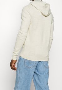 Felpa lavorata a maglia di un beige chiaro con cucitura sul retro e polsini a costine. Abbinata a jeans in denim blu chiaro, comodi e con tasche.