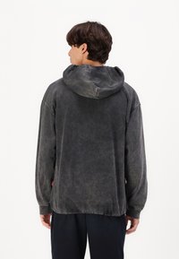 Sötét szürke, oversized kapucnis pulóver, puha, enyhén kopott textúrával, húzózsinórral ellátott kapucnival és hosszú ujjakkal. Minimális részletezéssel.