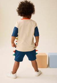 Light beige t-shirt with blue sleeves, colorful spikes along the back, paired with blue shorts, and white shoes.